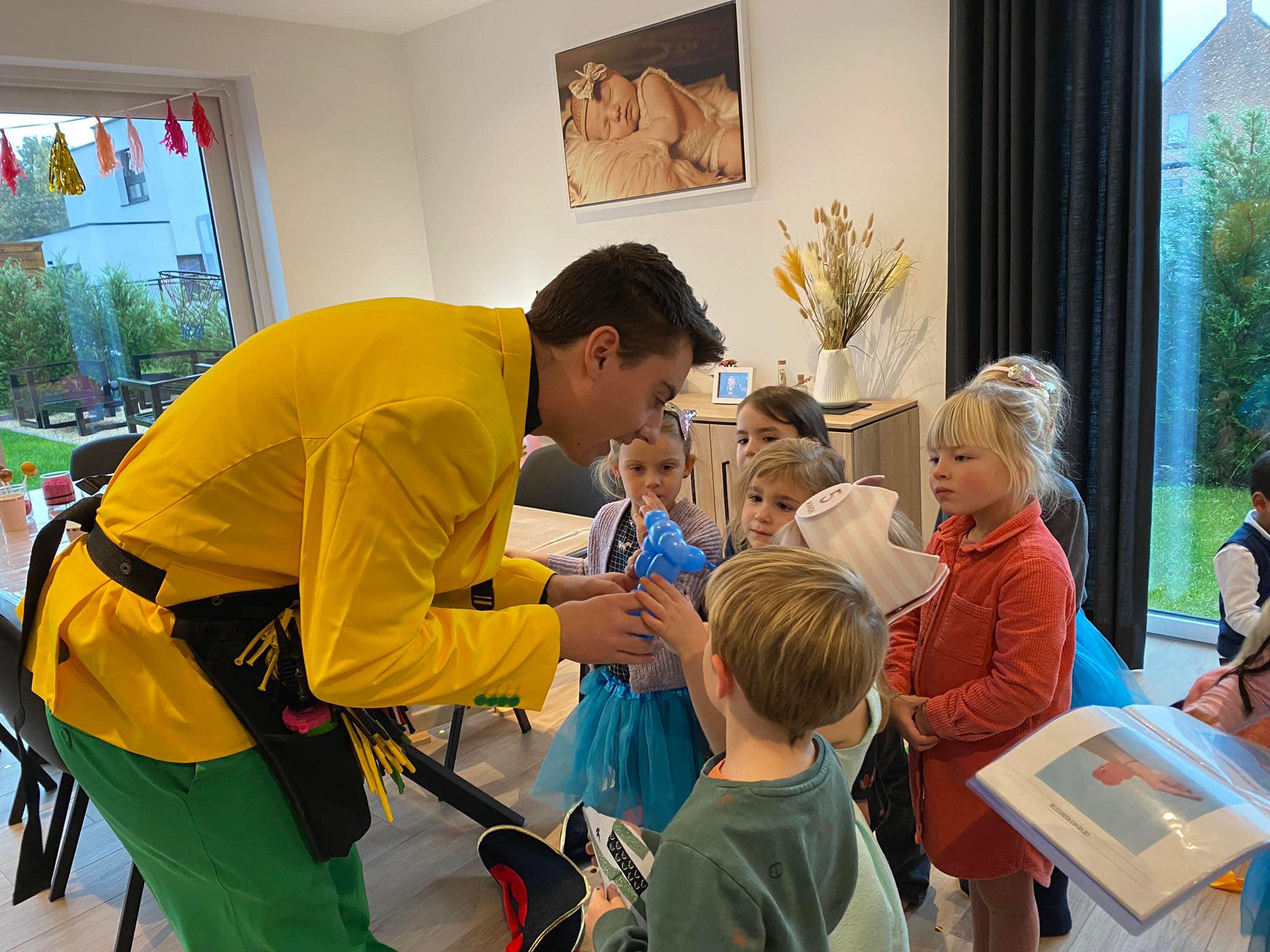 Een ballonplooier in een geel pak geeft een blauw ballondier aan een groep kinderen.