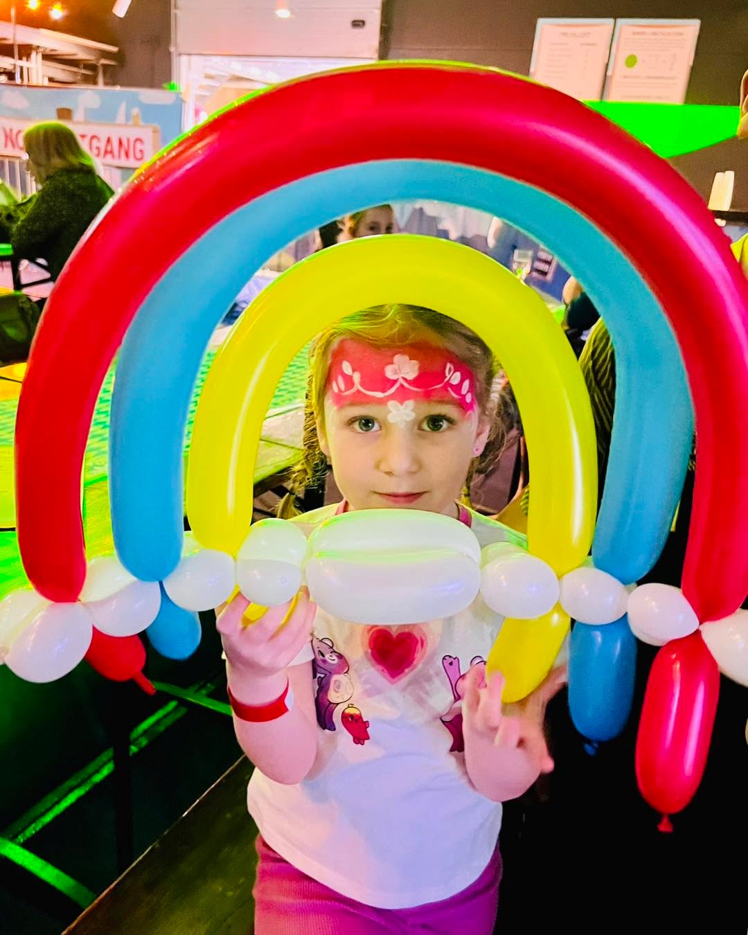Een meisje met roze bloemengrime kijkt door een grote ballonregenboog.