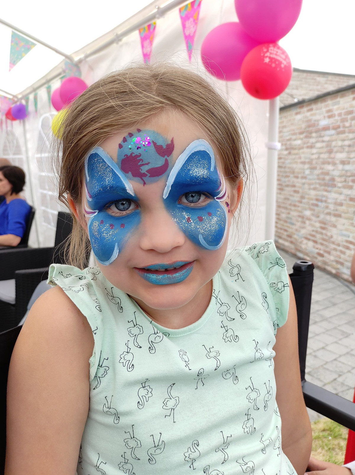 Een meisje met blauwe vlindergrime en glitter rond haar ogen.