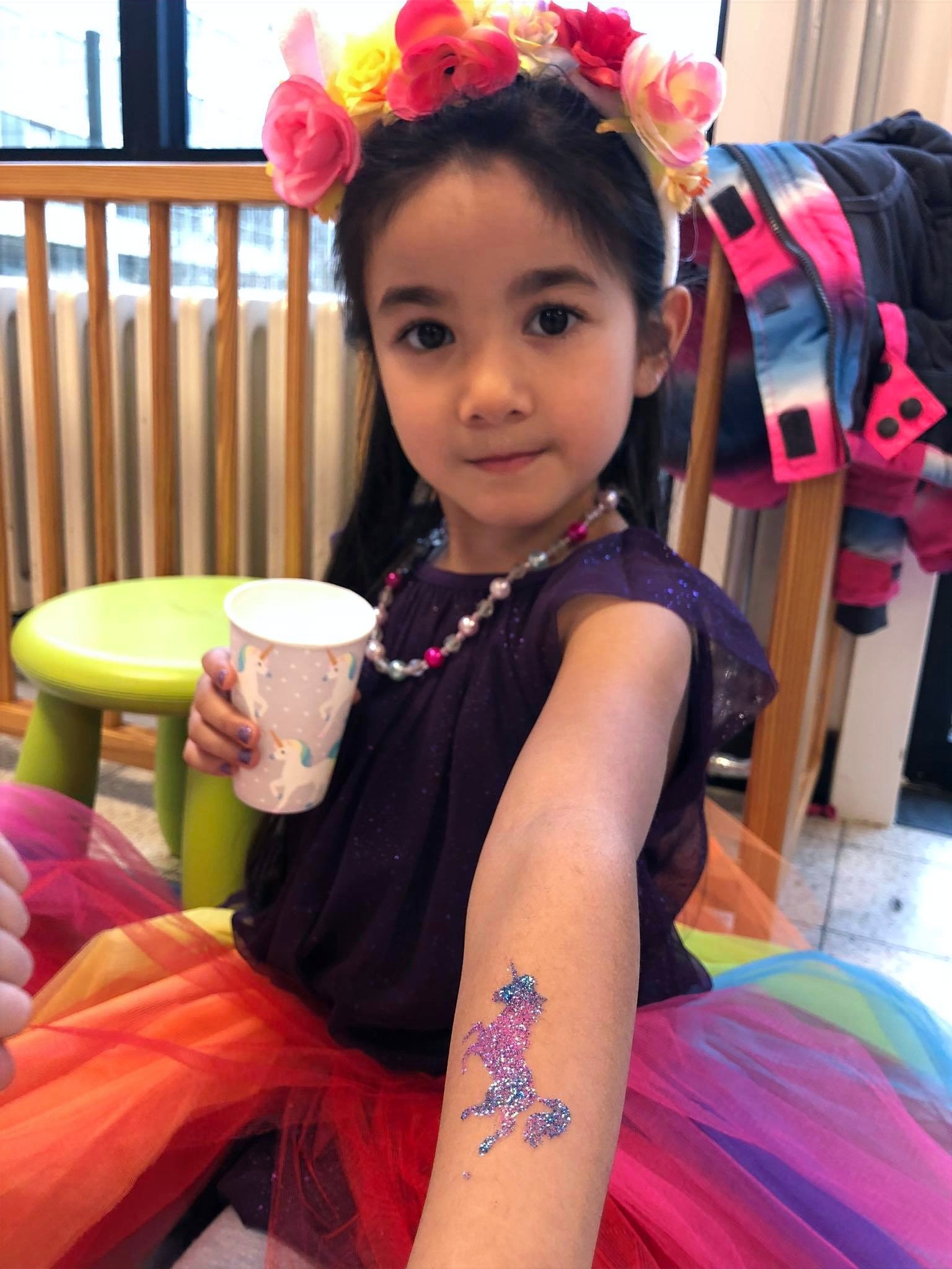 Een meisje met een bloemenkrans toont een glinsterende eenhoorntattoo op haar arm.