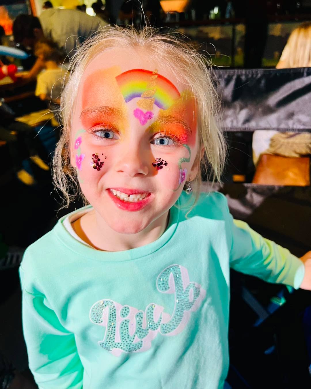 Een lachend meisje met eenhoorn- en regenbooggrime en glitter op haar wangen.