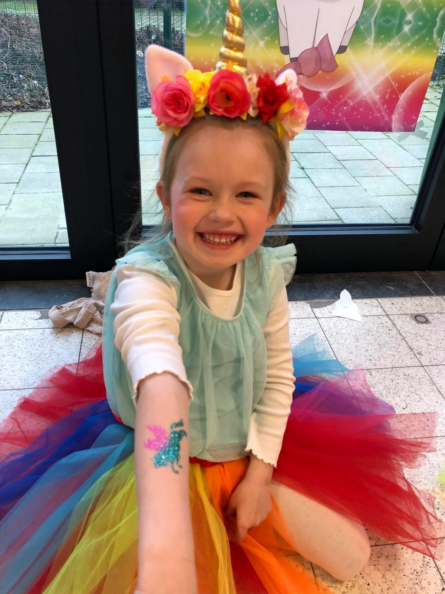 Een lachend meisje met een eenhoornkroon toont haar kleurrijke glittertattoo.