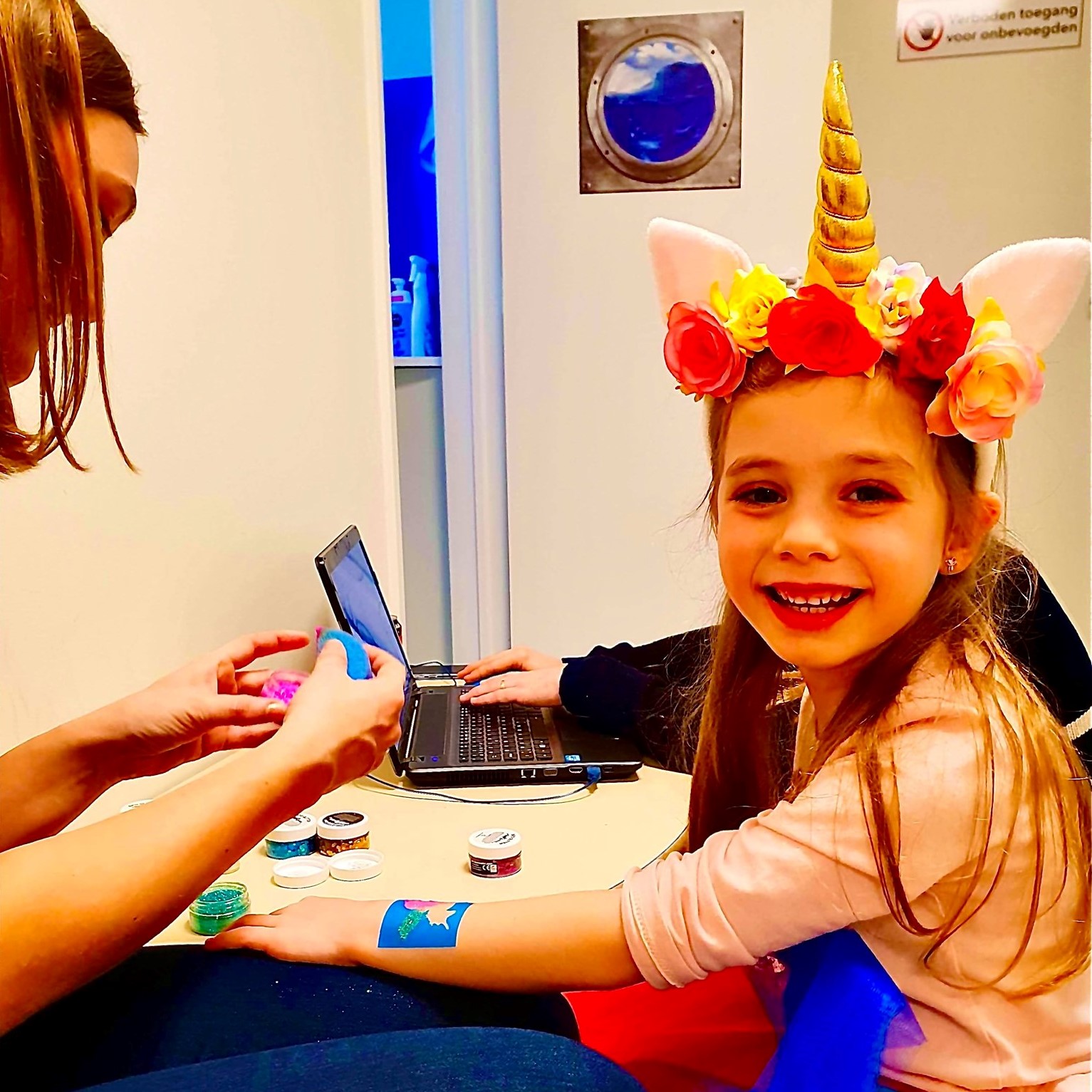 Een meisje kijkt lachend toe terwijl een glittertattoo op haar arm wordt aangebracht.