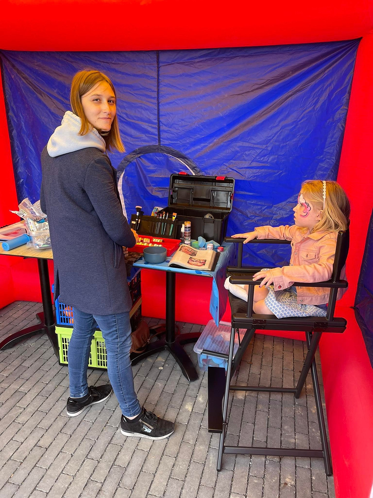 Een grimeuse schminkt een meisje in de grimeerstoel onder een tentje.