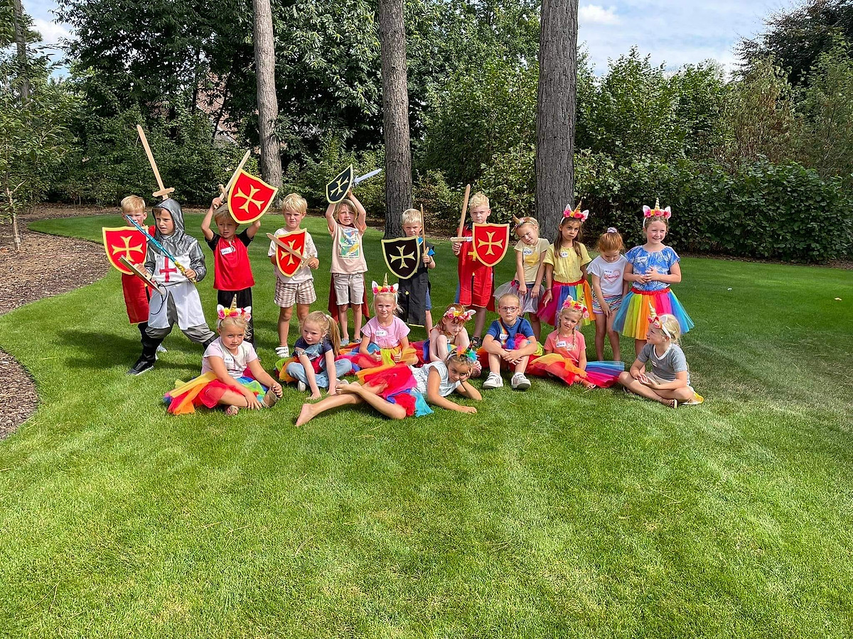 Een grote groep kinderen verkleed als ridders en eenhoorns poseert samen in het gras.
