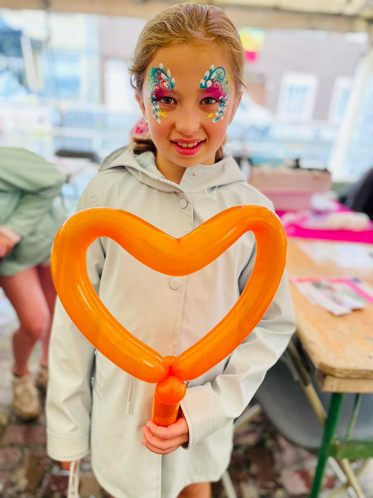 Een meisje met glittergrime houdt een groot oranje ballonhart vast.