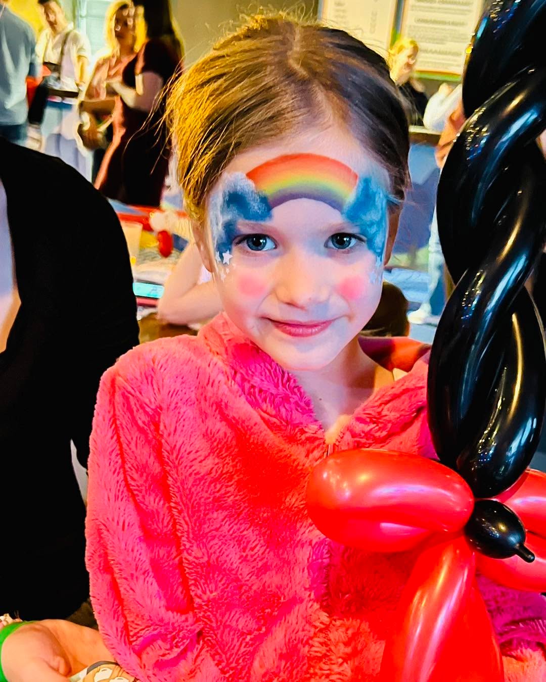 Een meisje met een regenboog en wolkjes op haar voorhoofd toont een rood-zwart ballonfiguur.