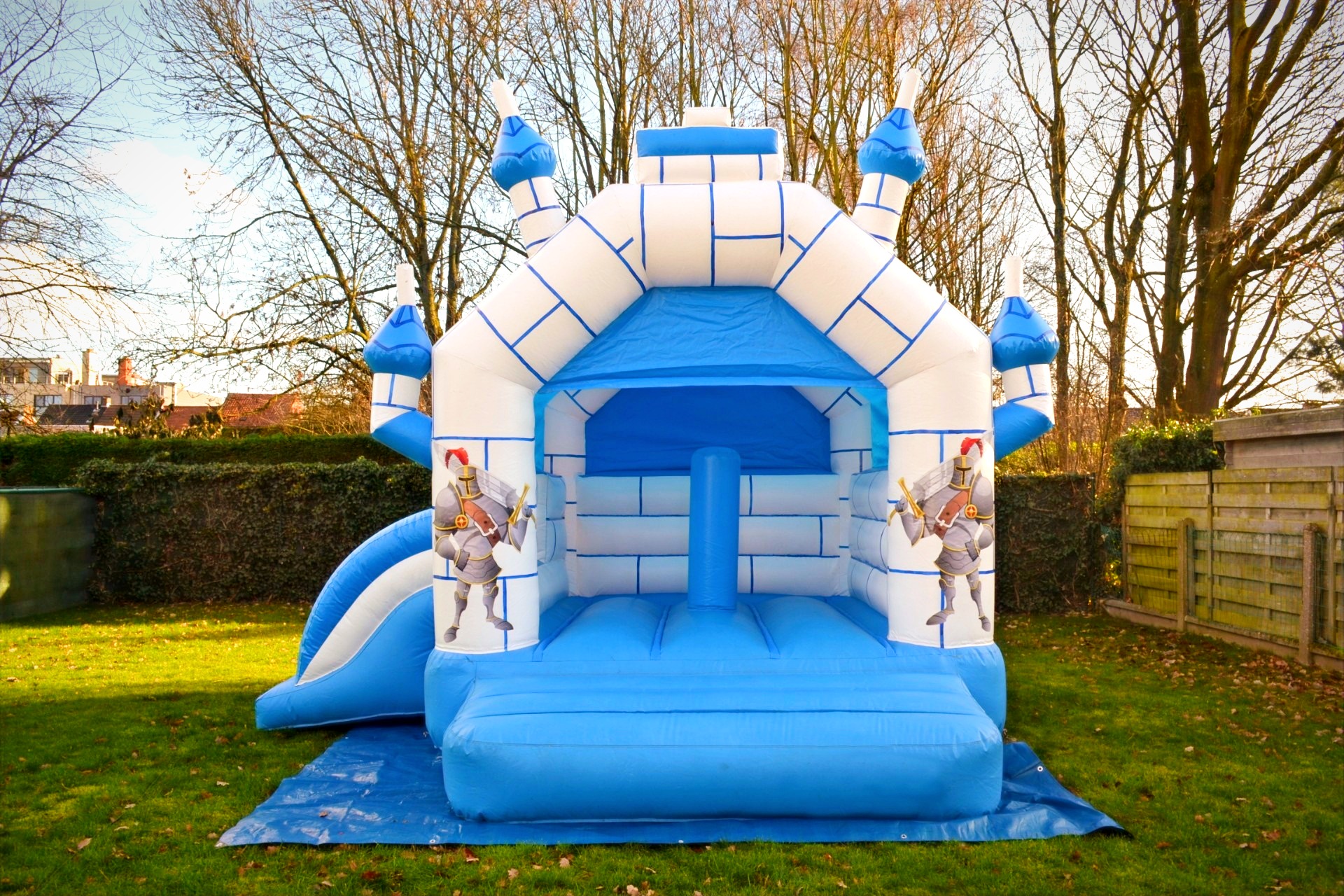 Springkasteel Ridder in een tuin, blauw met wit en met ridders op de torens. De glijbaan hangt aan de linkerkant. Foto 1 van 8.
