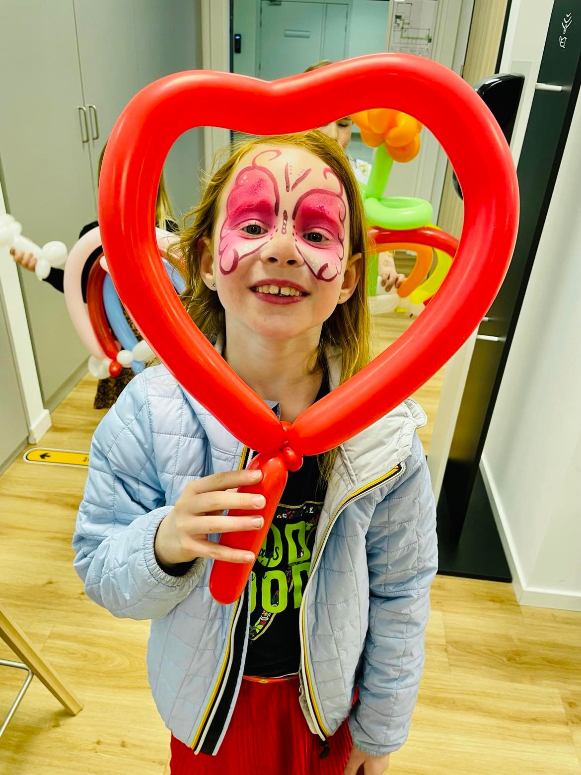 Een meisje met roze vlindergrime kijkt door een hartvormige rode ballon.